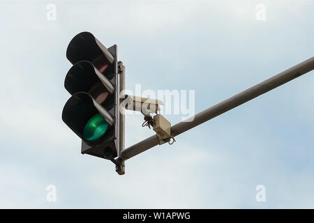 Outdoor surveillance camera and green traffic light both installed on a pole above a roadway, against a blue sky. Modern automatic traffic control.