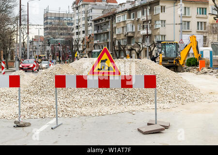 The sign of road works on red white barrier in front of a pile of gravel on a city street. Construction and repair of asphalt roads in a town. City im Stock Photo