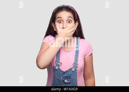 Portrait of shocked brunette young girl in pink t-shirt and blue denim overalls standing, covering her mouth and looking at camera with big eyes. indo Stock Photo