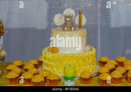 A two tier birthday cake surrounded by yellow cupcakes on a table. Stock Photo