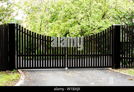 Dark wooden driveway property entrance gates set in timber picket fence with garden shrubs and trees in background Stock Photo