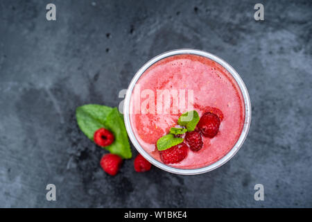 Top view of a raspberry smoothie glass set on a wooden boards, decorated with drinking straw, three raspberries and mint leaves. Selective focus. Stock Photo