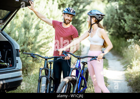 Young couple preparing for the bicycle riding, standing near the car trunk on the forest road during the summer vacations Stock Photo