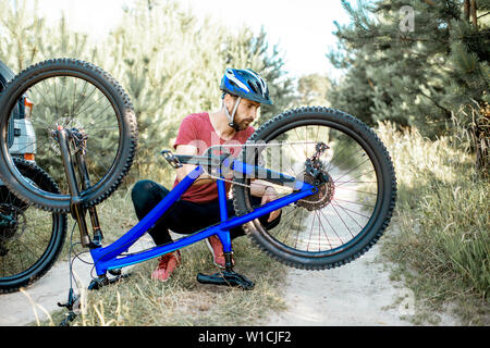 Young man in sportswear repairing bicycle on the forest road during the summer time Stock Photo