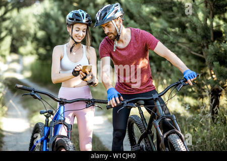 Young man and woman cyclists looking on the smartphone, choosing the way for riding bicycles, while standing on the forest road during the summer time Stock Photo