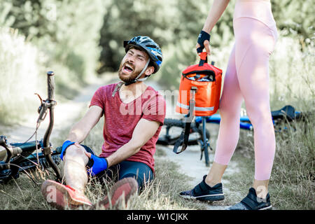 Woman giving first aid to a man with knee injury while cycling on the mountain bicycles on the forest road during the summer time Stock Photo