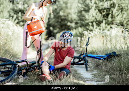 Woman giving first aid to a man with knee injury while cycling on the mountain bicycles on the forest road during the summer time Stock Photo