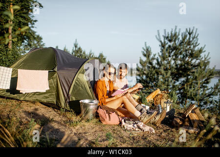 Young and cheerful couple having a picnic at the campsite while traveling in the forest near the lake during the sunset Stock Photo