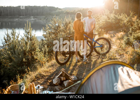 Young couple standing with mountain bicycles at the campsite, traveling in the forest near the lake on the sunset Stock Photo