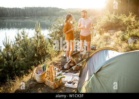 Young couple standing at the campsite, talking and enjoying hot drinks while traveling in the mountains near the lake on the sunset Stock Photo