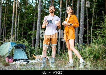 Young couple standing with hot drinks at the camping in the forest, enjoying their summer trip during the sunset Stock Photo