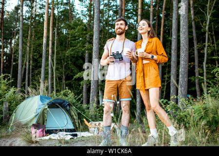 Young couple standing with hot drinks at the camping in the forest, enjoying their summer trip during the sunset Stock Photo