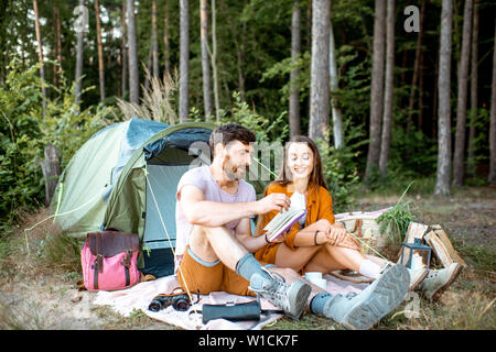 Young and cheerful couple sitting with book near the tent, enjoying the summer vacations in the forest during the sunset Stock Photo