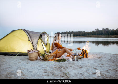 Young woman sitting at the fireplace, cooking sausages, having a picnic at the campsite on the beach in the evening Stock Photo