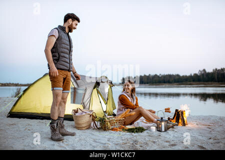 Man and woman at the campsite on the beach, man carrying a bucket with fresh caught fish, woman cooking sausages on the fireplace Stock Photo