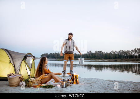 Man and woman at the campsite on the beach, man carrying a bucket with fresh caught fish, woman cooking sausages on the fireplace Stock Photo