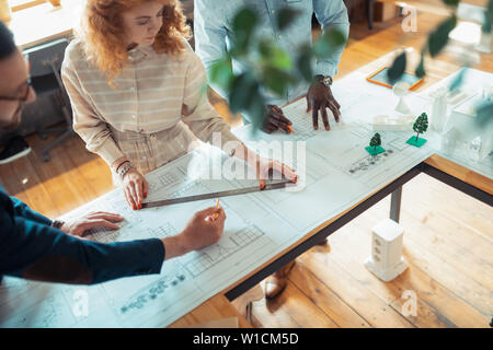 Woman making sketches. Curly red-haired experienced woman making sketches standing near colleagues Stock Photo