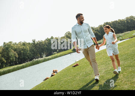 Enjoying great time with father. Happy young father and daughter holding hands and smiling while walking near the river. Love and family concept Stock Photo