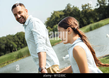 Enjoying great time with father. Back view of happy young father and daughter holding hands and smiling while walking near the river. Love and family concept Stock Photo