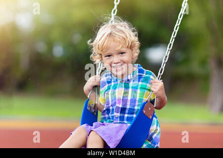 Child playing on outdoor playground in rain. Kids play on school or kindergarten yard. Active kid on colorful swing. Healthy summer activity for child Stock Photo