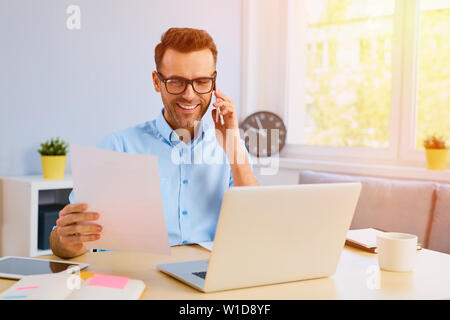 Man talking on the phone from his home office, holding some papers Stock Photo