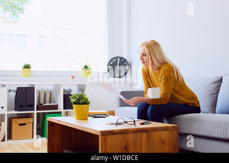 Beautiful young woman sitting on sofa drinking coffee and reading document Stock Photo