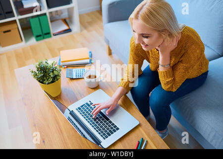 Gorgeous smiling young woman sitting on sofa and using laptop Stock Photo