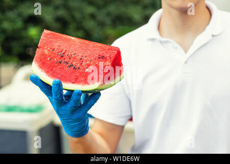 Cook in rubber gloves holding in hand and offering big piece of fresh tasty juicy sliced watermelon for hotel guests at tropical resort outdoors Stock Photo