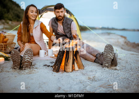 Young and cheerful couple cooking sausages at the fireplace, having a picnic at the campsite on the beach in the evening Stock Photo