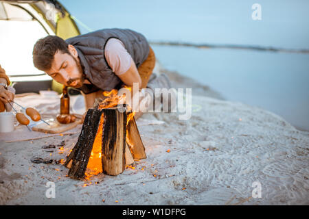 Young and cheerful couple cooking sausages at the fireplace, having a picnic at the campsite on the beach in the evening Stock Photo