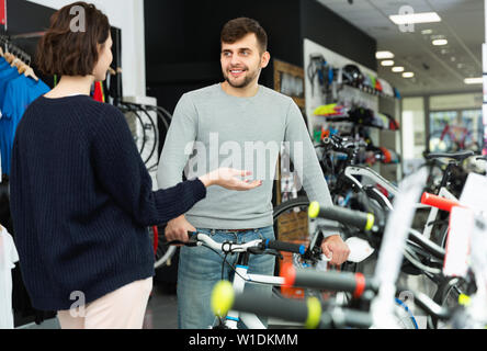 Young cheerful positive smiling man seller demonstrating bicycle to woman client in store Stock Photo