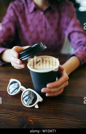 Female hands holding a cup of cappuccino with thick foam in black takeaway paper cup. Coffee take away. Stock Photo