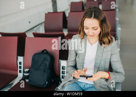 Young attractive girl at the airport waiting for her departure. Scrolling smartphone. Coffee cup and backpack beside. Stock Photo