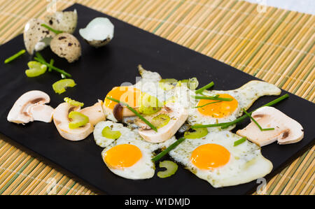 Delicious broken quail eggs with green onion and sliced champignons on black serving board Stock Photo