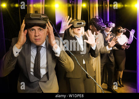 Toned image of shocked businessman taking off VR goggles on background of colleagues. Concept of mixing illusion and reality in business Stock Photo