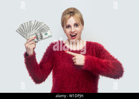 Happy amazed attractive young businesswoman in red blouse standing, holding many dollars, pointing finger, looking at camera with open mouse and big e Stock Photo