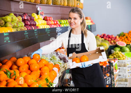 Positive female seller putting fresh goods on shelves in greengrocery Stock Photo