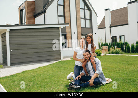 Happy heteroseksual family sitting on grass near their new house. Family sitting on lawn, buying new big home . Stock Photo