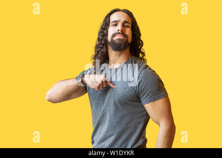 Portrait of proud bearded young man with long curly hair in grey tshirt standing, pointing himself and looking at camera with confident serious face. Stock Photo