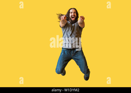 Portrait of surprised bearded young man with long curly hair in grey tshirt jumping, pointing and looking at camera with amazed happy funny face. indo Stock Photo