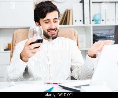 Cheerful brunette man holding glass of red wine and having video chat at office Stock Photo