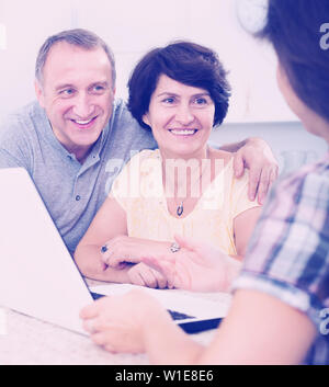 Attentive positive mature couple looking at woman showing documents at her laptop indoors. Focus on both mature persons Stock Photo