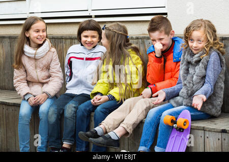 Group of happy positive children sitting on bench and sharing secrets outdoors Stock Photo