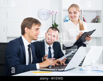 Two happy business male assistants working using laptops and woman with cardboard taking notes in firm office Stock Photo