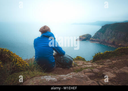 Young hiker man sitting with his backpack on the edge of a cliff to contemplate the scenic coastline landscape. Travel, lifestyle, outdoors concept Stock Photo