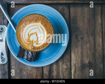 Piccolo Latte coffee topping with flower art from milk in small glass with a piece of homemade brownies cake on blue ceramic plate on wooden tray on w Stock Photo