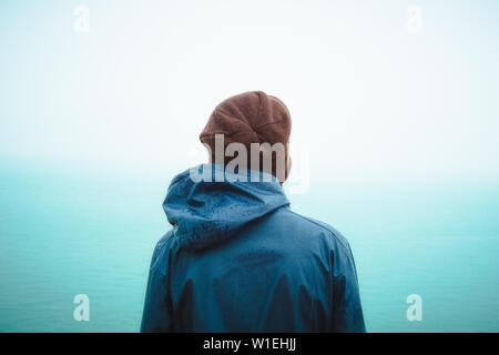 Rear or back view of a young woman wearing a blue rain coat and brown wooly hat standing alone and watching the ocean on a rainy and moody day. Outdoo Stock Photo