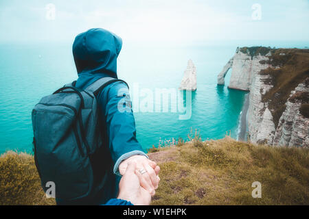 Follow me. Young woman in blue raincoat taking hand of her boyfriend walking to the edge of the cliff to watch a scenic seascape, Etretat, France. Stock Photo