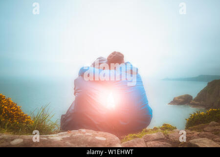 Rear or back view of young lovers sitting against each other and hugging on a cliff edge looking at a scenic seascape with light burst. Love, romance, Stock Photo