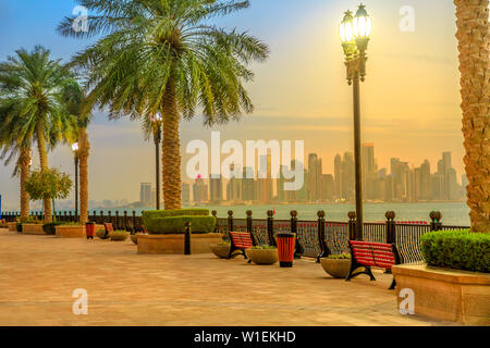 Benches and palm trees along marina walkway in Porto Arabia at the Pearl-Qatar, with skyscrapers of West Bay skyline, Doha, Qatar Stock Photo